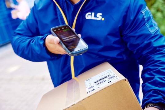 GLS delivery worker in a blue uniform using a handheld delivery terminal while holding a package.