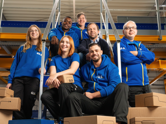 Équipe de collaborateurs GLS en tenue professionnelle dans un entrepôt logistique, entourée de colis, illustrant les opportunités de carrière et les métiers du transport et de la logistique.