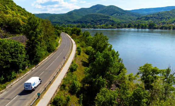 Camion blanc GLS roulant sur une route sinueuse en bordure d'une forêt dense et d'un lac, vue aérienne.