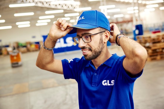 Collaborateur GLS en uniforme bleu, souriant dans un entrepôt logistique, en train d'ajuster sa casquette.