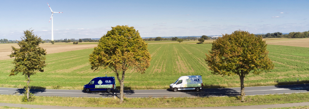 Deux camionnettes de livraison GLS sur une route de campagne, avec champs verts et éoliennes à l'horizon.