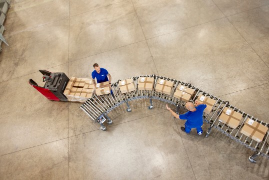 Opérateurs au travail dans un entrepôt logistique GLS, le long d'un convoyeur à rouleaux chargé de cartons, illustrant la chaîne opérationnelle et la performance logistique du groupe.