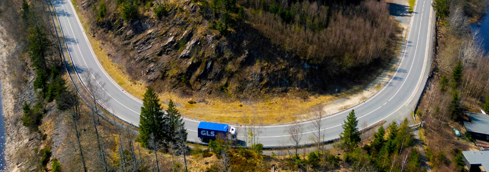 Camion semi-remorque GLS bleu sur une route de montagne en lacet, en milieu naturel.